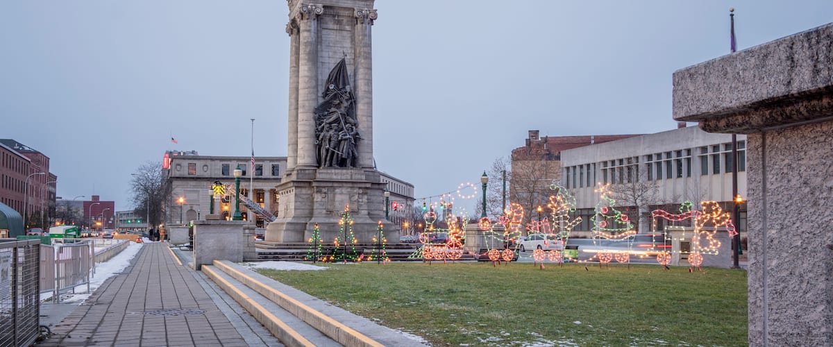 A monument with a statue, surrounded by a paved walkway and decorated with holiday lights.