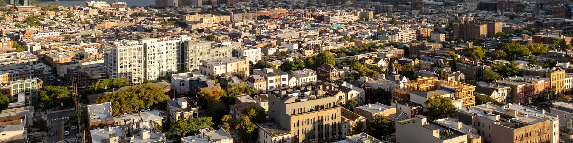 Wide aerial view capturing the neighborhoods of Hoboken and Jersey City, with the iconic New York City skyline stretching across the horizon in the background. Overcast skies add depth