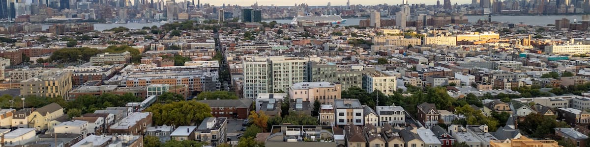 Wide aerial view capturing the neighborhoods of Hoboken and Jersey City, with the iconic New York City skyline stretching across the horizon in the background. Overcast skies add depth