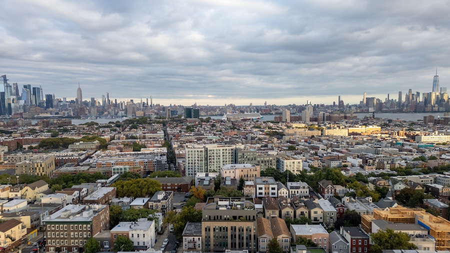 Wide aerial view capturing the neighborhoods of Hoboken and Jersey City, with the iconic New York City skyline stretching across the horizon in the background. Overcast skies add depth