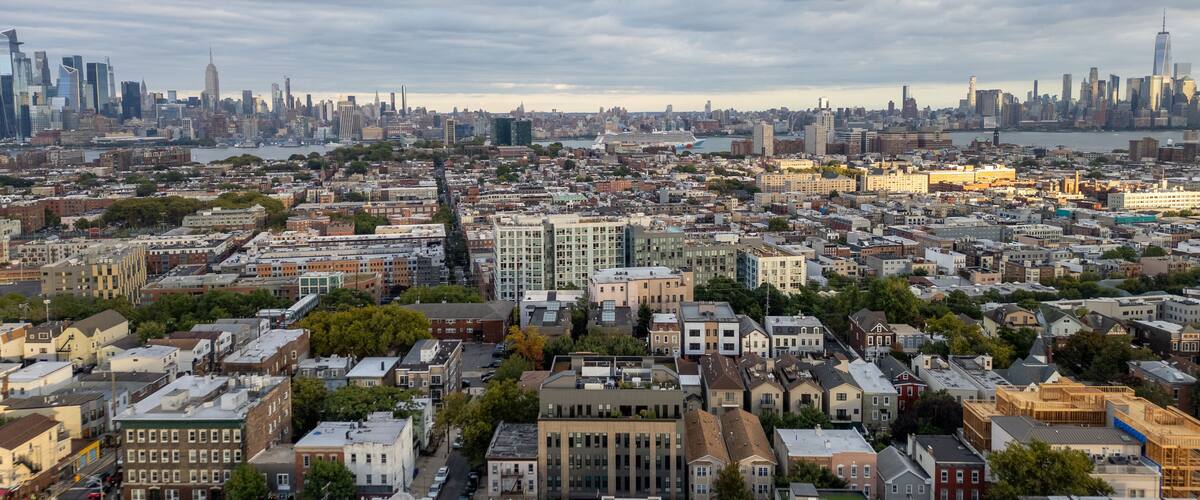 Wide aerial view capturing the neighborhoods of Hoboken and Jersey City, with the iconic New York City skyline stretching across the horizon in the background. Overcast skies add depth