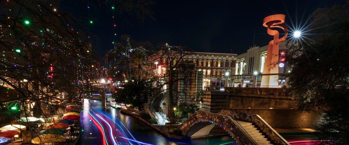 Ein beleuchtetes Stadtbild bei Nacht, mit Straßen, einer Brücke und einer großen Skulptur.