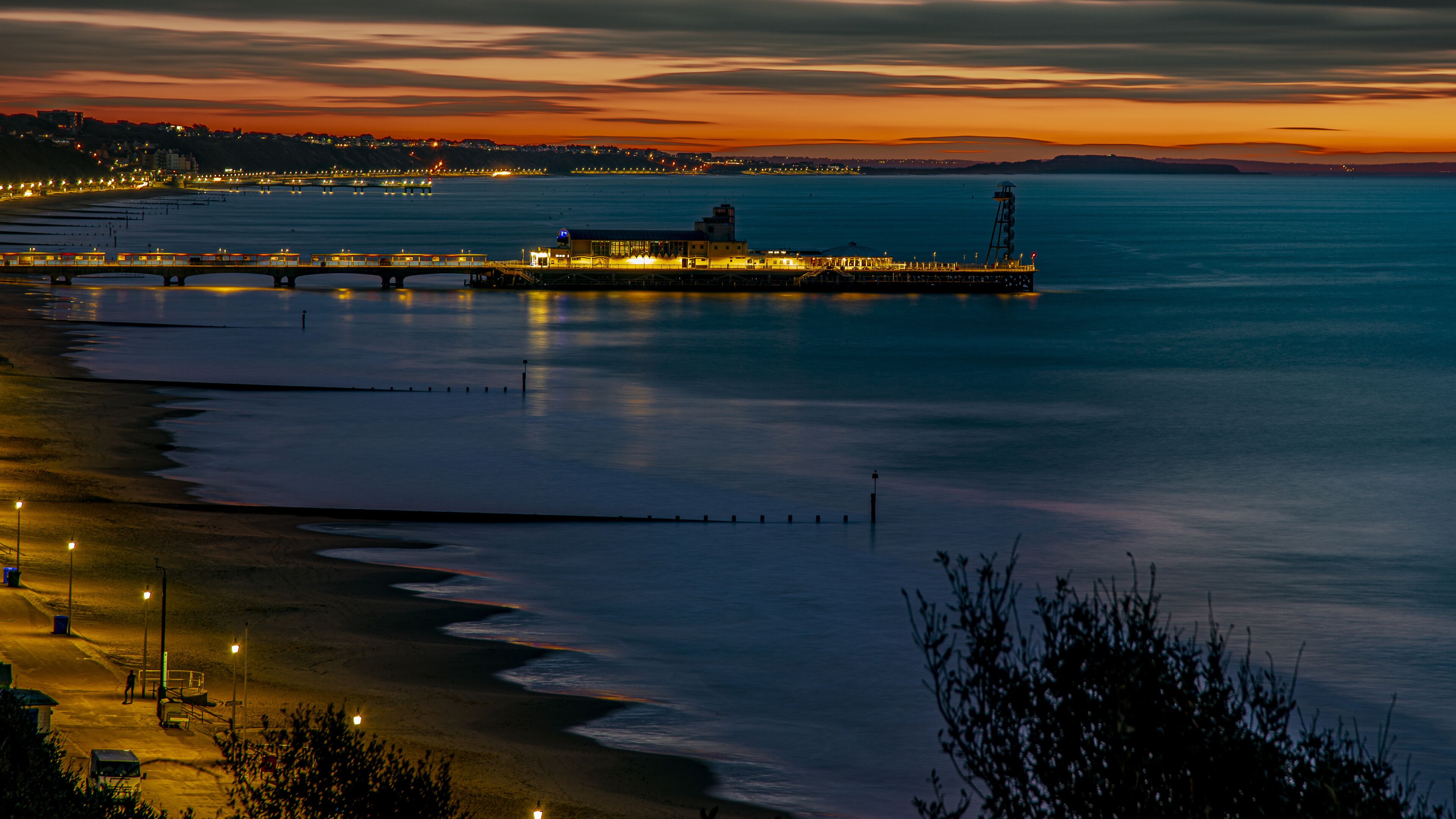 A coastal sunset with a pier extending into the water, a beach with a fence, and a distant city skyline.