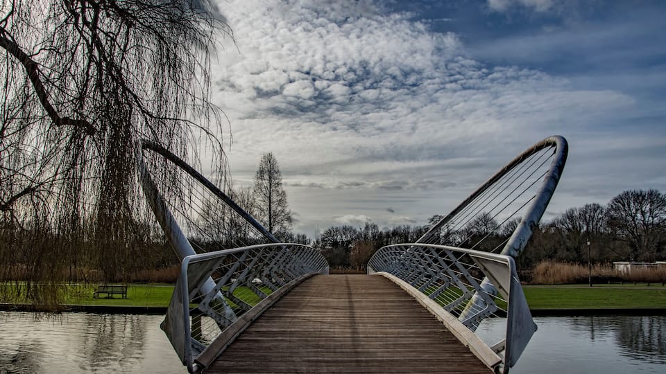 A wooden bridge with metal railings over a calm body of water, surrounded by trees and a grassy area.