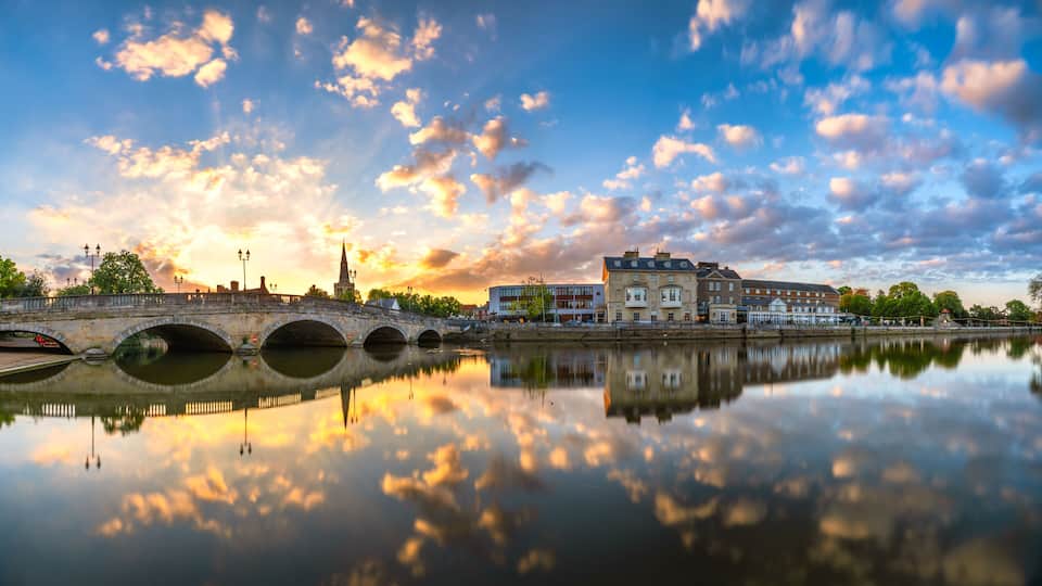 A river with a stone bridge, a church spire, and buildings reflecting in the water.