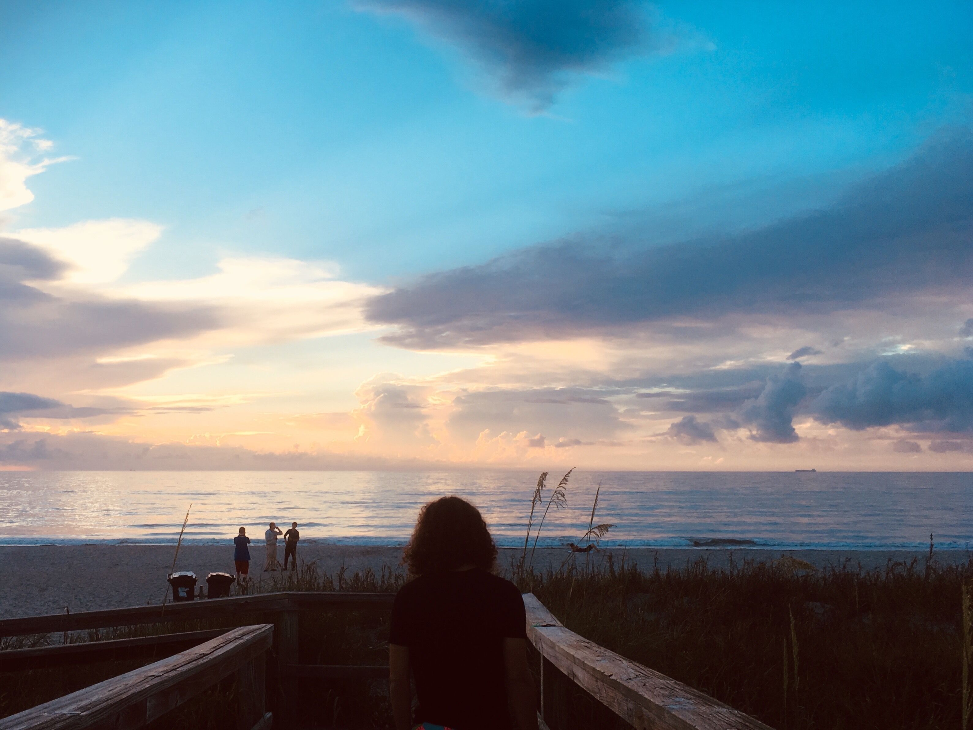 A beach with a wooden boardwalk, people standing and walking, and a sunset over the ocean.