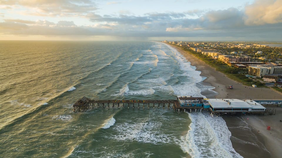 A coastal city with a pier extending into the ocean, waves crashing onto the shore, and buildings lining the beach.
