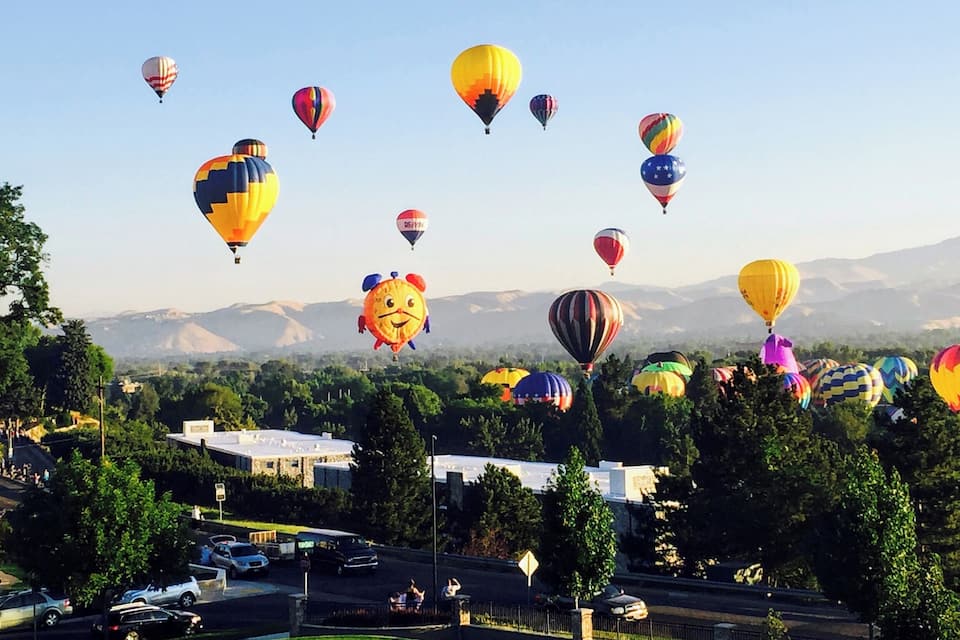 A cityscape with a large number of colorful hot air balloons in the sky, a highway with cars, and a mountainous background.