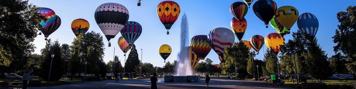 A variety of colorful hot air balloons in a park with a fountain.