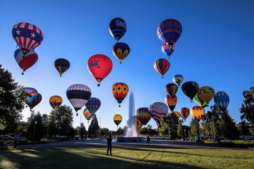 A variety of colorful hot air balloons in a park with a fountain.