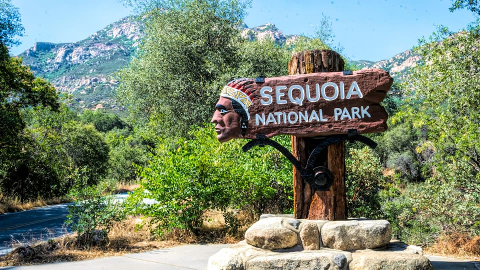 A wooden sign with 'SEQUOIA NATIONAL PARK' written on it, surrounded by greenery and mountains in the background.