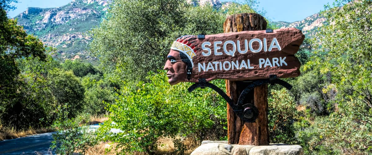 A wooden sign with 'SEQUOIA NATIONAL PARK' written on it, surrounded by greenery and mountains in the background.