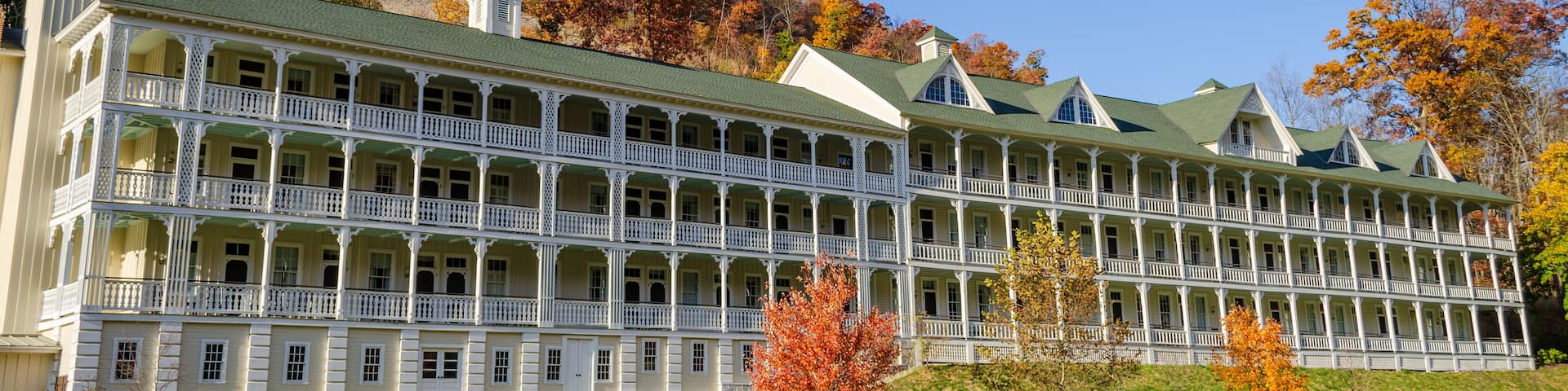A large, multi-story building with a green roof and white balconies, surrounded by autumn-colored trees and a clear blue sky.
