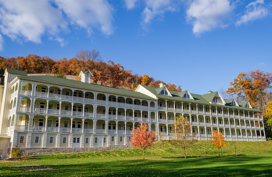 A large, multi-story building with a green roof and white balconies, surrounded by autumn-colored trees and a clear blue sky.