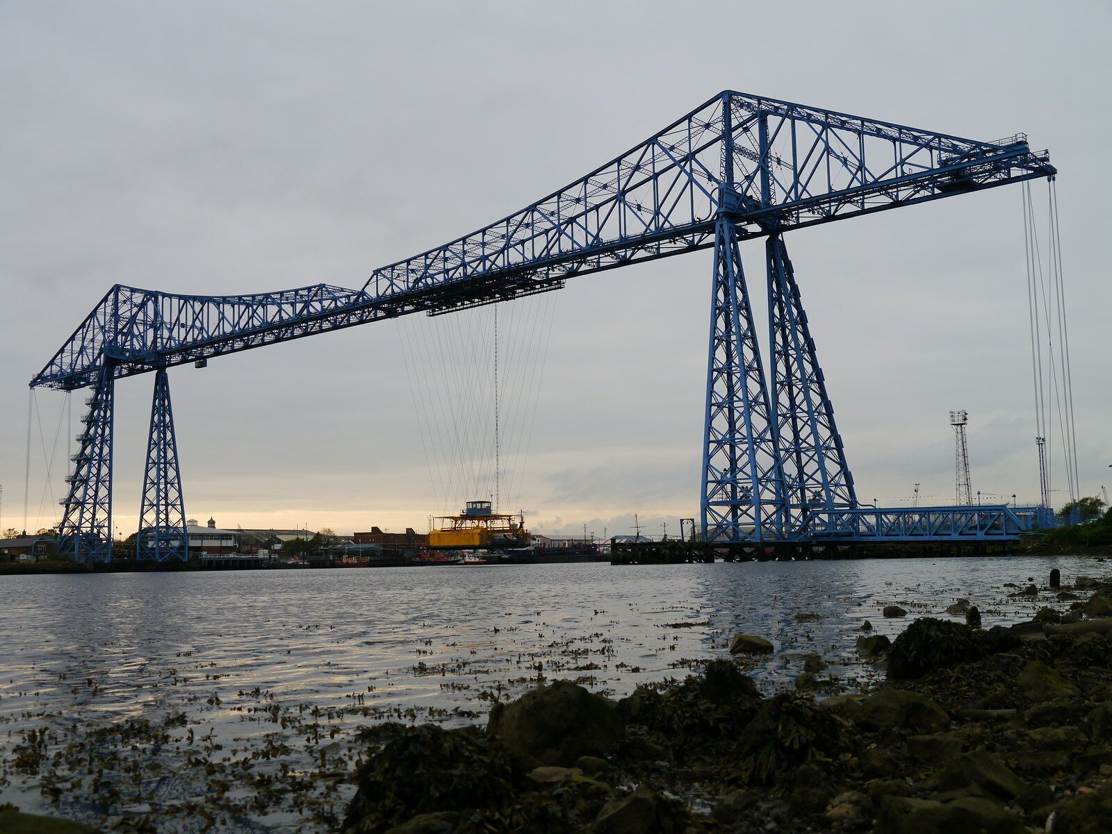 A large blue crane structure over water with a rocky shoreline in the foreground.