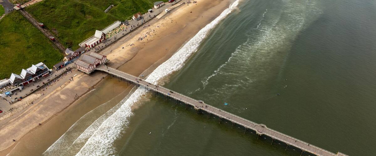 A pier extending into the sea with a beach and buildings nearby.