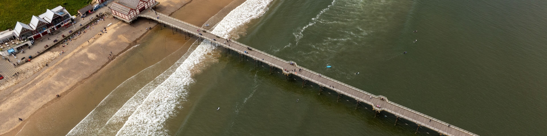 A pier extending into the sea with a beach and buildings nearby.