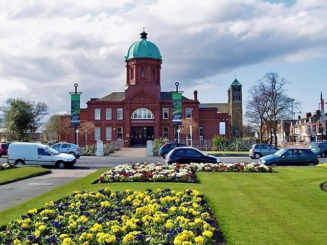 A historic building with a green dome and a clock tower, surrounded by well-maintained flower beds and parked cars.