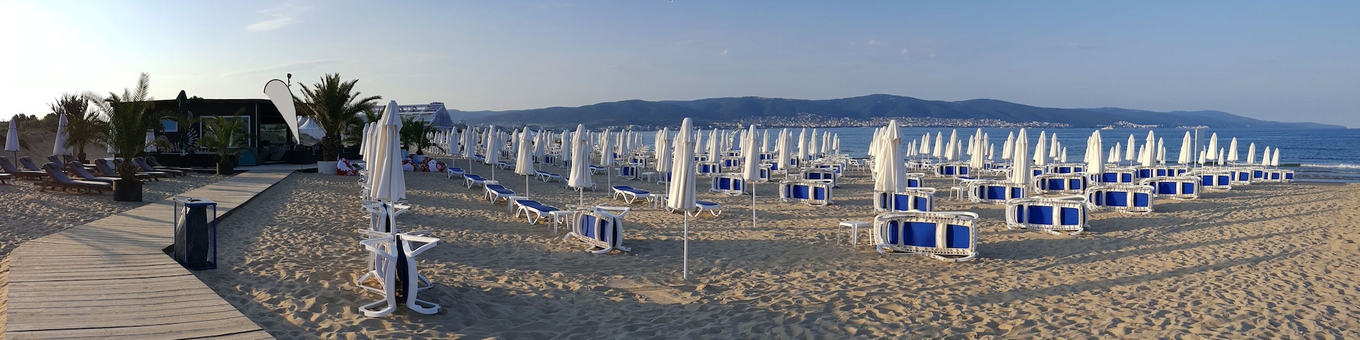 A beach with rows of sun loungers and umbrellas, a wooden boardwalk, and palm trees.