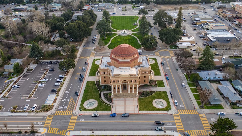 An aerial view of a city with a prominent octagonal building with a red roof, surrounded by a well-maintained park and intersecting roads.