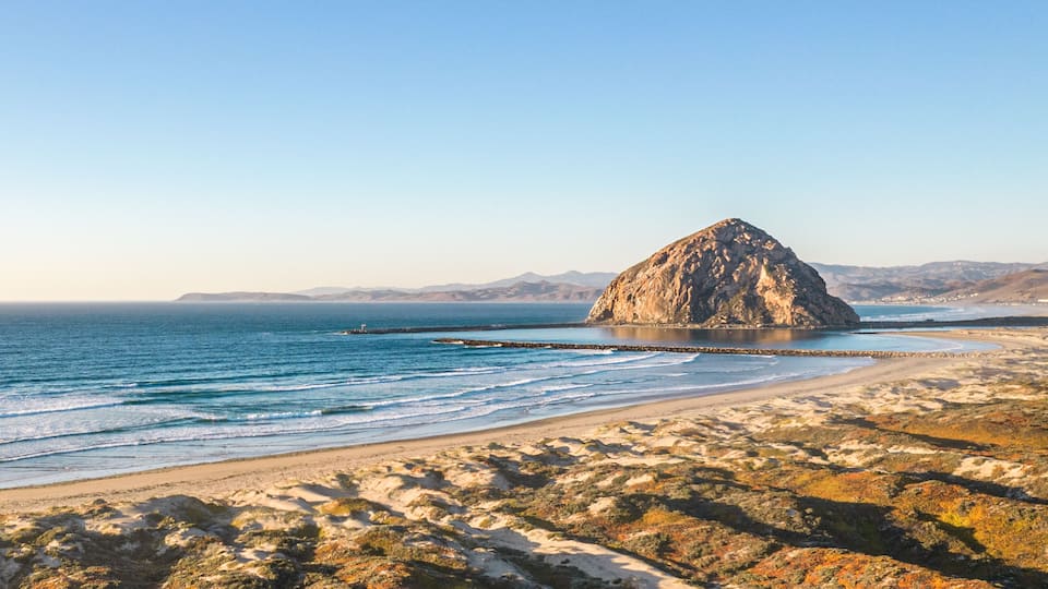 A rocky island in the ocean with a sandy beach and distant mountains.