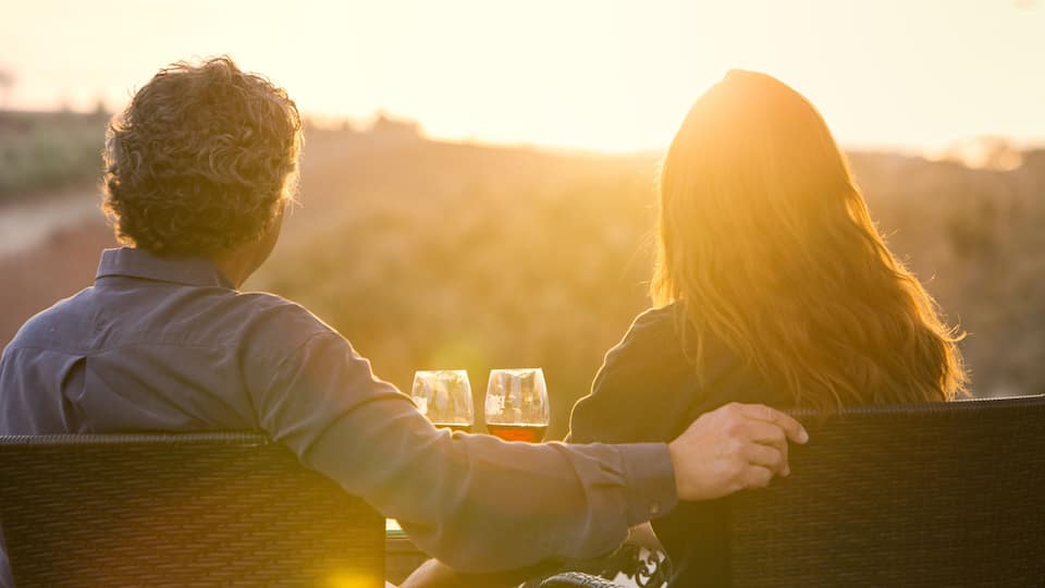Two people sitting on a bench facing away from the camera, with a sunset in the background.