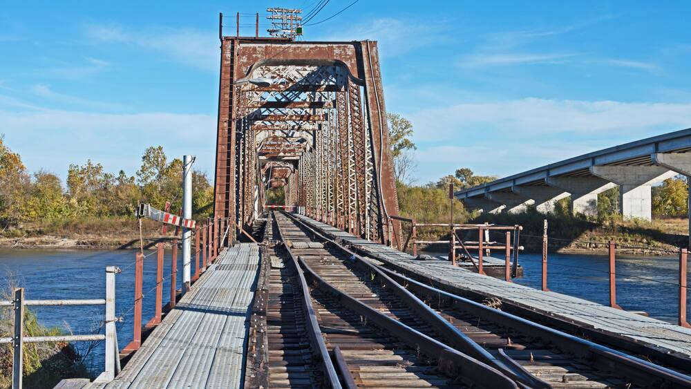 Un pont ferroviaire rouillĂ©, avec des voies ferrĂ©es qui sâenfoncent dans le lointain, entourĂ© dâarbres et bordĂ© par une Ă©tendue dâeau.