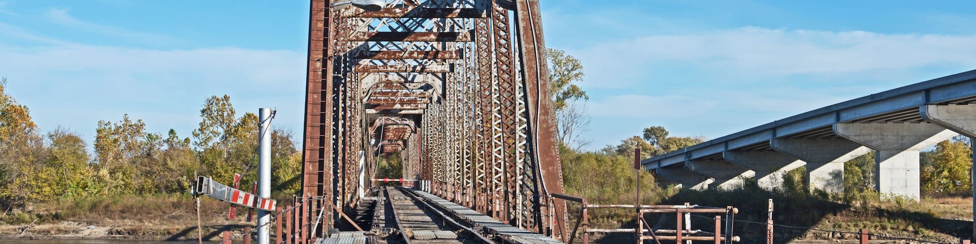 A rusted railway bridge with tracks leading into the distance, surrounded by trees and a body of water.