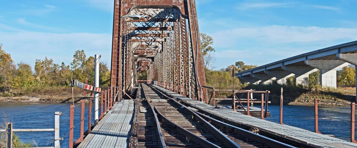 A rusted railway bridge with tracks leading into the distance, surrounded by trees and a body of water.