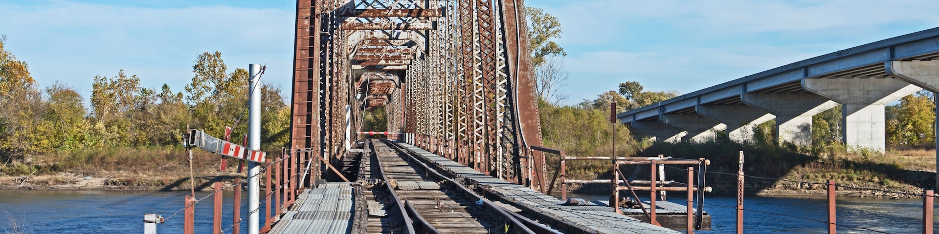 A rusted railway bridge with tracks leading into the distance, surrounded by trees and a body of water.