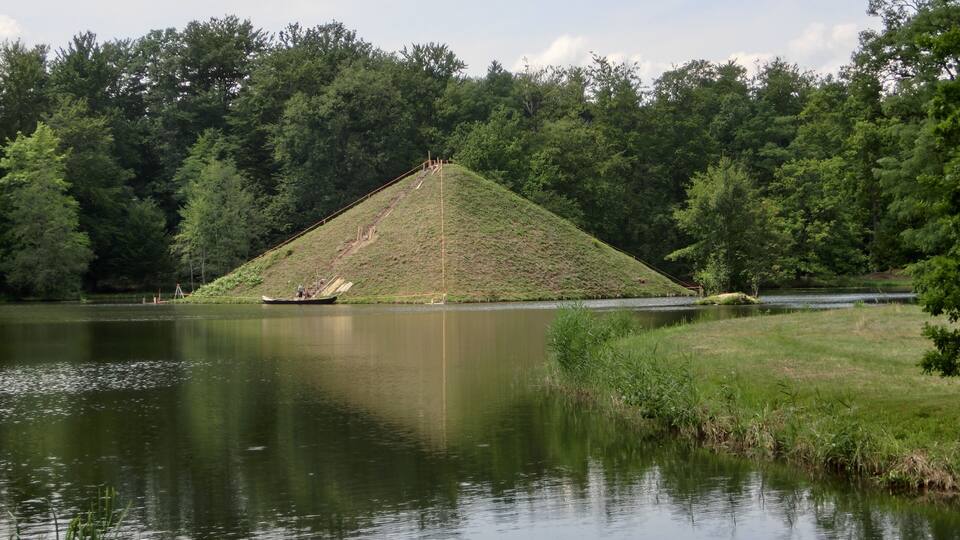 A calm lake with a grassy hill and dense trees in the background.