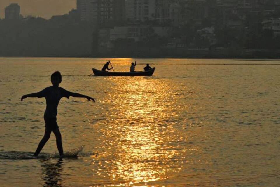 Un atardecer sobre el horizonte de una ciudad, con edificios silueteados contra el cielo, una persona vadeando el agua y un bote con dos personas a lo lejos.