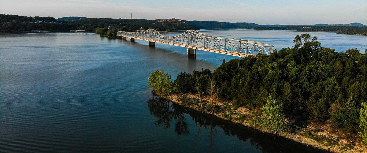 A steel bridge spanning a calm river, surrounded by dense forest and distant hills.