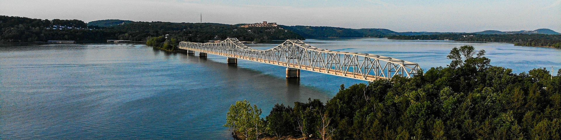 A steel bridge spanning a calm river, surrounded by dense forest and distant hills.