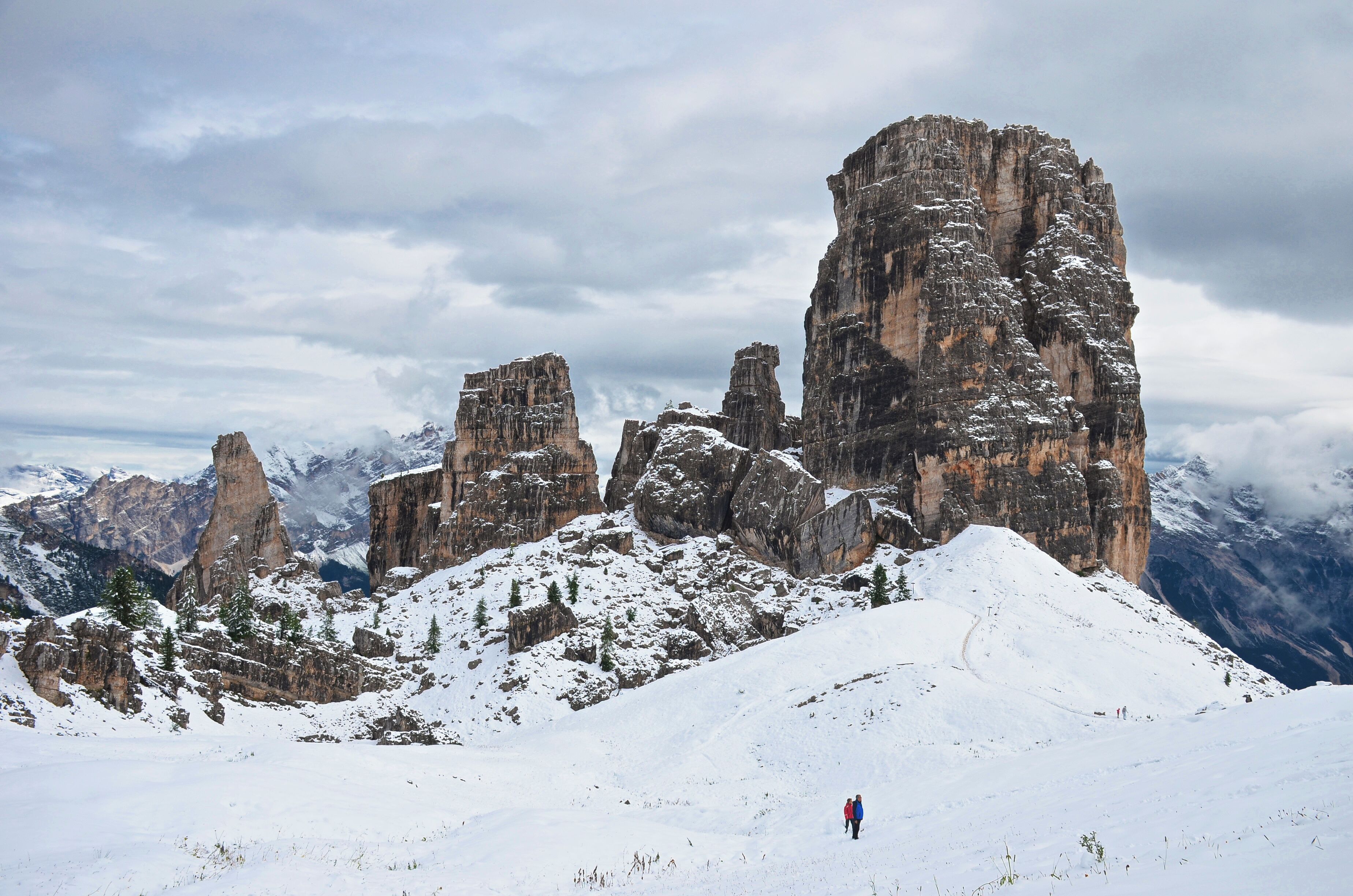 A snow-covered mountain landscape with prominent rock formations and a hiker in the foreground.