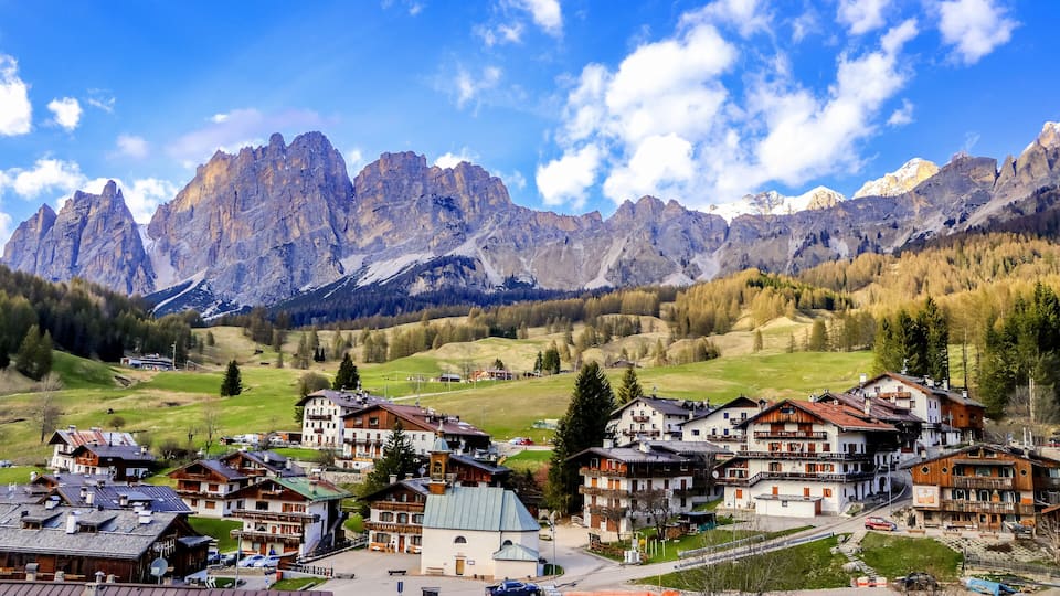 A mountain village with traditional houses, a road, and a valley with trees and mountains in the background.