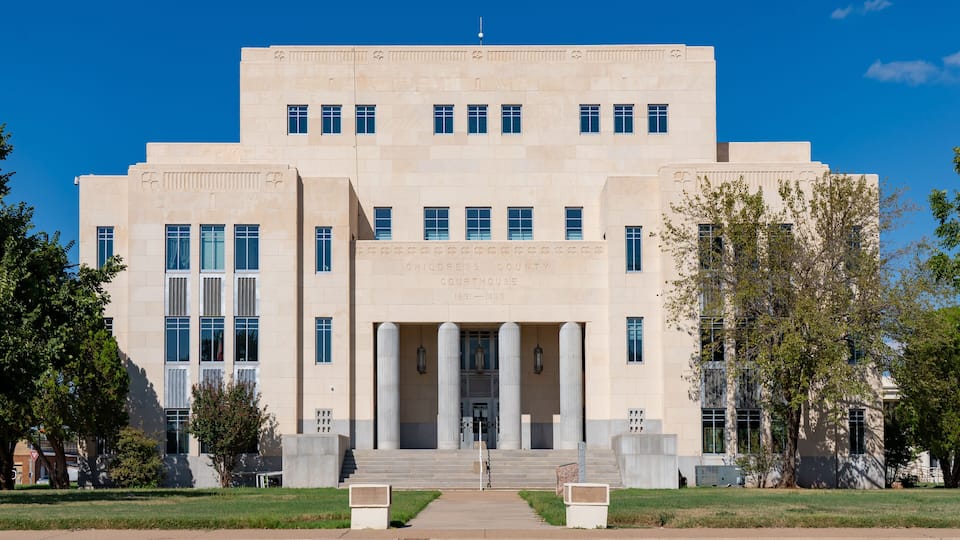 A large, beige courthouse with a central entrance flanked by columns, surrounded by a well-maintained lawn and trees.