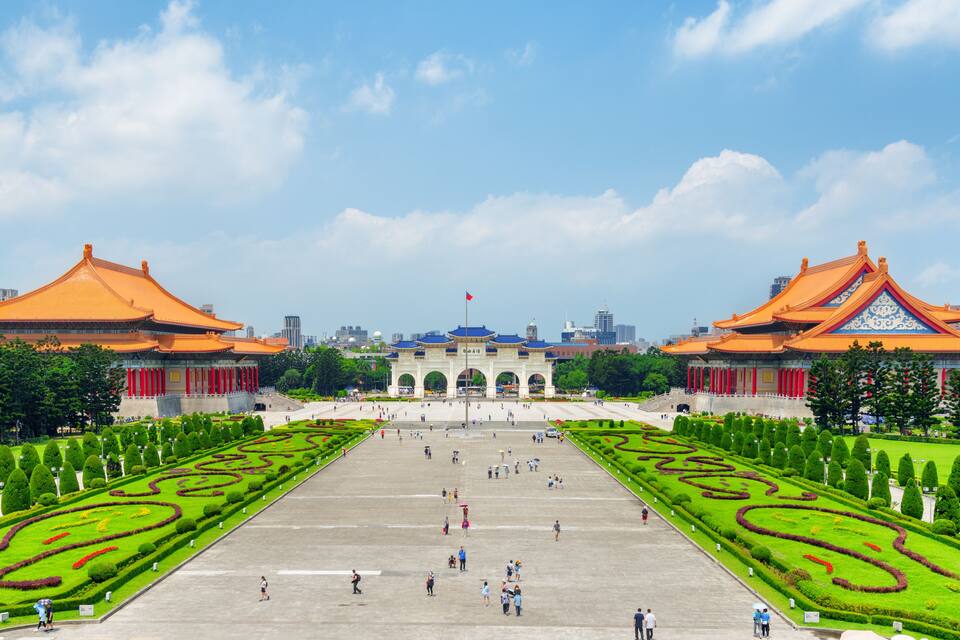 A large open plaza with two traditional buildings, a flagpole, and a well-maintained garden with geometric patterns.