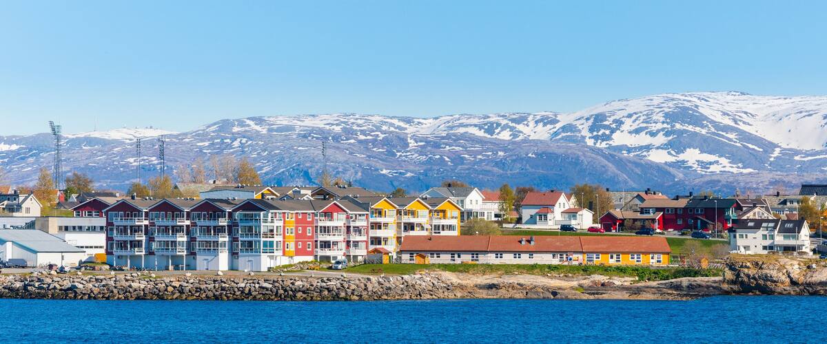 A coastal town with colorful houses, a rocky shoreline, and snow-capped mountains in the background.