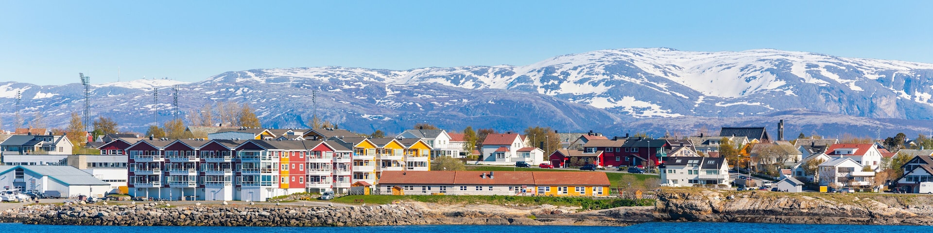 A coastal town with colorful houses, a rocky shoreline, and snow-capped mountains in the background.