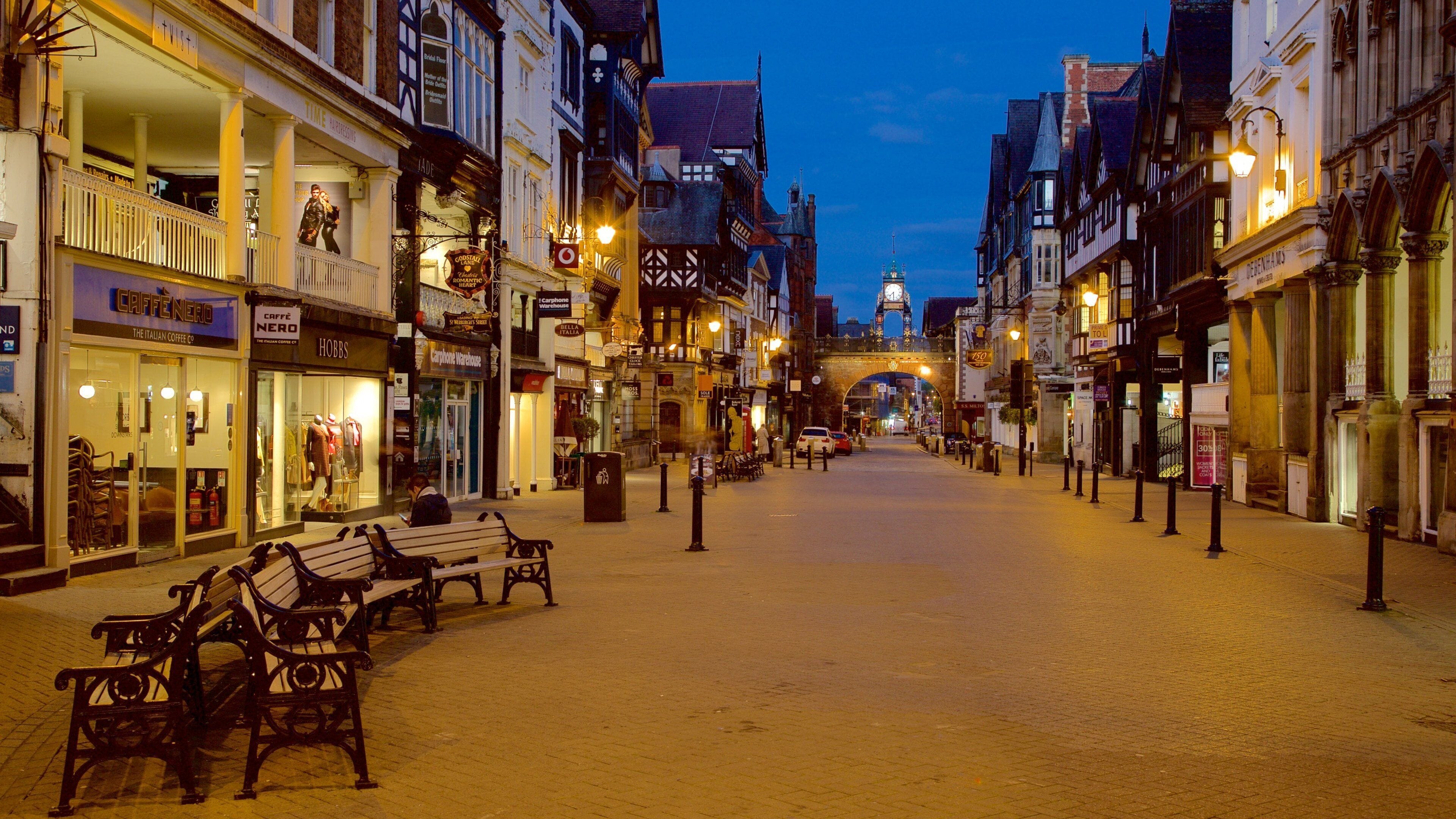 A cobblestone street lined with historic buildings, benches, and a clock tower in the background.