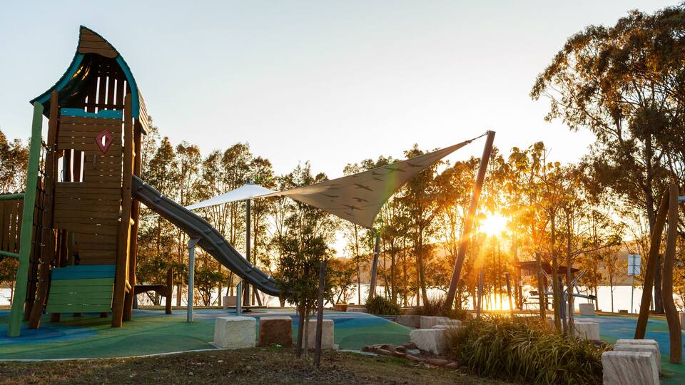 A playground with a slide and climbing structures, surrounded by trees and a body of water.