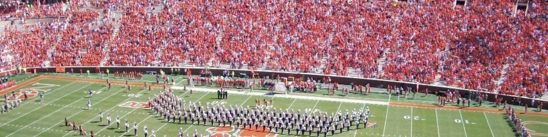 A football stadium filled with spectators, a marching band formation, and a football field with yard lines.