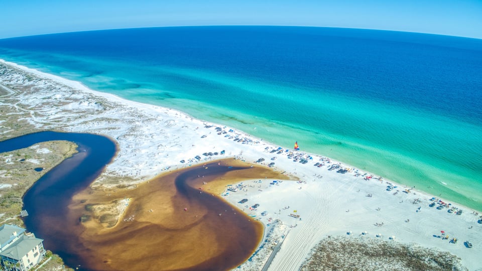 An aerial view of a beach with clear turquoise waters, a sandy shore, and a large body of brown water.