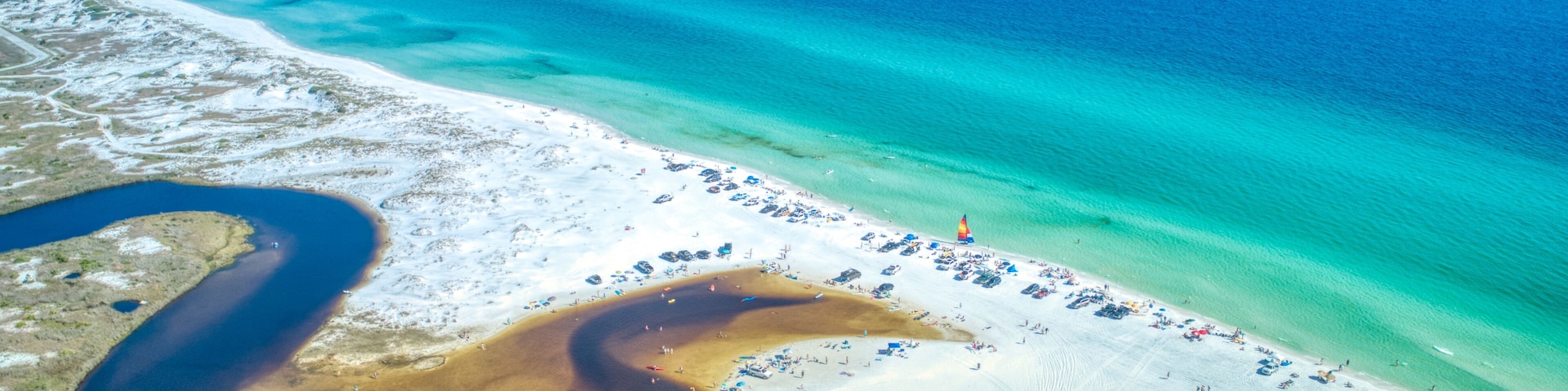 An aerial view of a beach with clear turquoise waters, a sandy shore, and a large body of brown water.