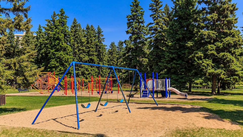A playground with swings and slides, surrounded by trees and a clear sky.