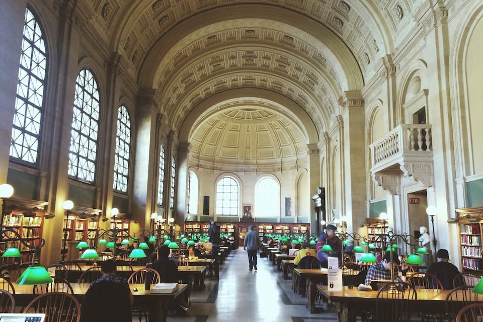 Una biblioteca amplia con techos altos, ventanas arqueadas y lámparas de lectura verdes.