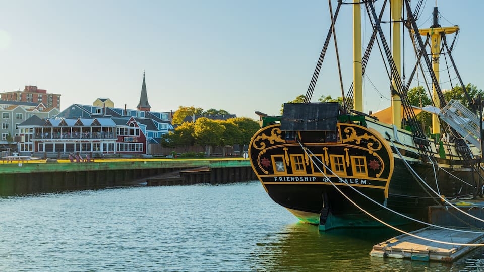 A historic ship named Friendship of Salem docked at a pier with a waterfront building and a church in the background.