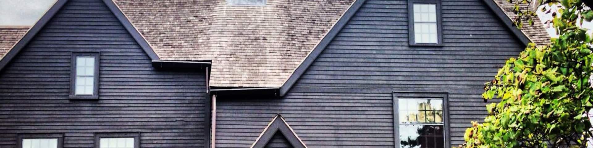 Une maison en bois sombre, avec un toit triangulaire, une cheminée en briques et une allée gravillonnée.