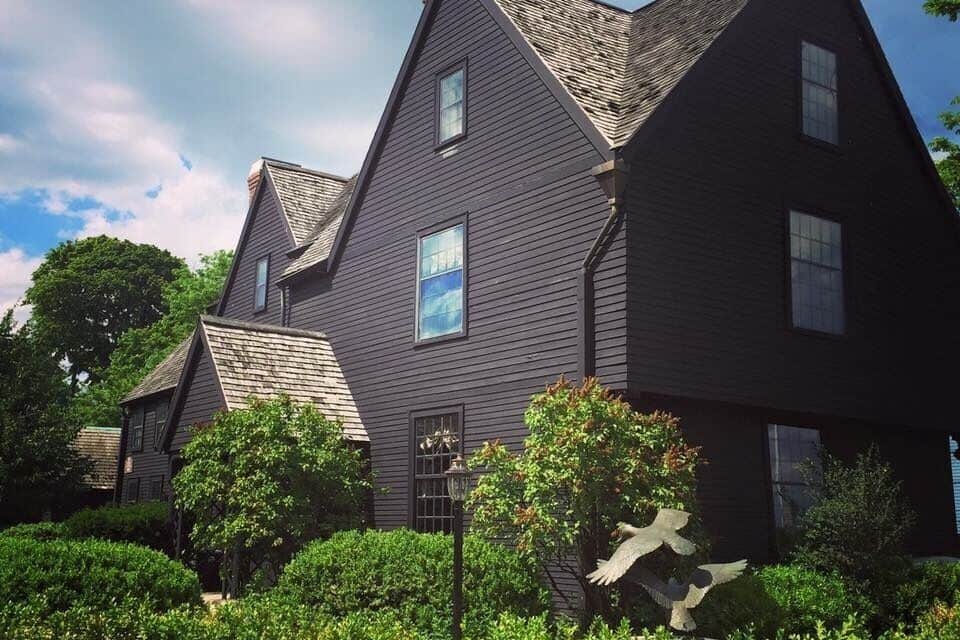 Une maison aux couleurs sombres, dotée de plusieurs toits à pignons et d’un jardin bien entretenu, agrémenté de divers arbustes et d’une sculpture d’oiseau.